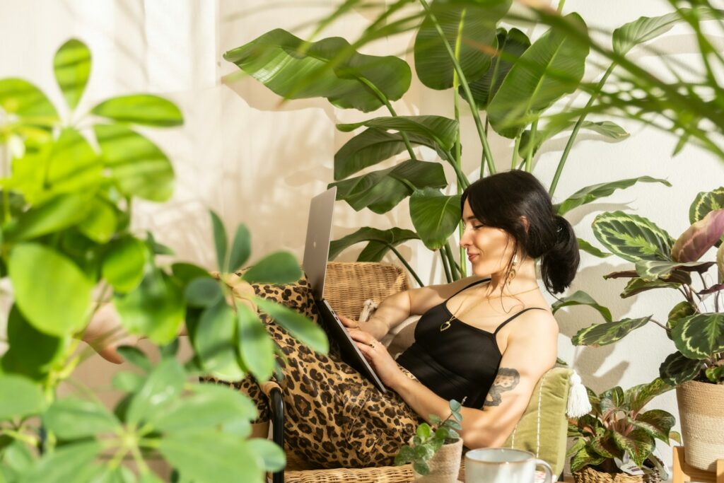 Donna rilassata seduta in casa tra piante e luce naturale mentre lavora al computer, esempio di routine di conforto quotidiana che aiuta a ridurre lo stress e ritrovare calma.