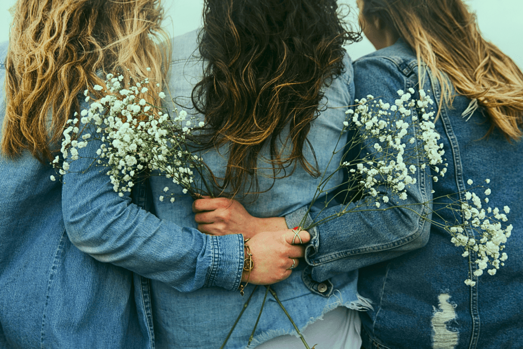Tre amiche abbracciate con piccoli fiori bianchi, immagine che rappresenta l’empatia tra donne e il conforto nelle relazioni femminili.