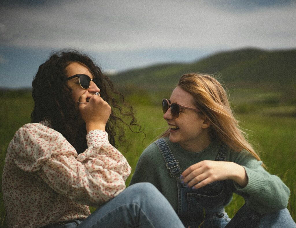 Due amiche sedute all’aperto ridono e si ascoltano, simbolo di una donna di conforto presente prima dei consigli.